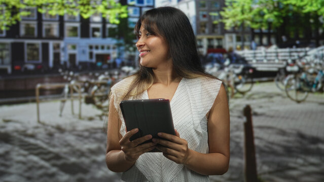 Latina woman holding tablet with hands visible on street by canal and bicycles; serenity exploration.