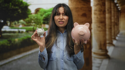 Young latina woman holds large and small piggybank in her hands on old town building street...