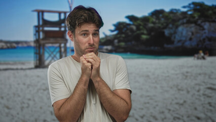 Man with clasped hands near mouth on sandy beach by lifeguard tower pleading expression and hunched shoulders, light tee visible; anxiety pleading.