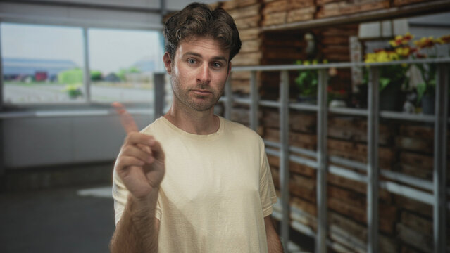 Man points finger forward in building aisle near shelving and crates, indoor setting with potted plants and window view; caution warning.