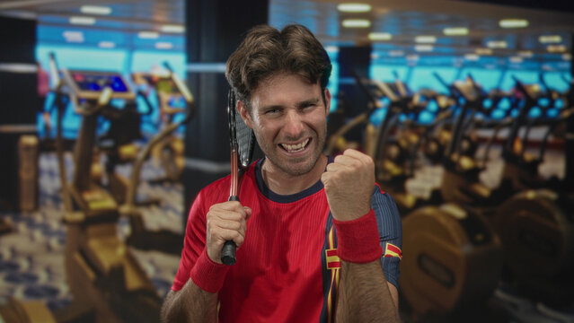 Man gripping tennis racket over shoulder and pumping clenched fist in gymnasium with treadmills and exercise bikes nearby; victory determination.