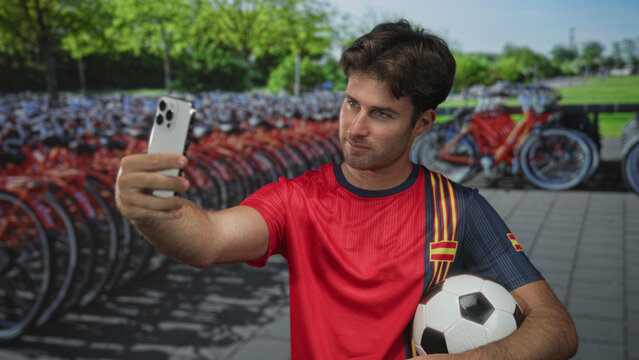 Man taking selfie on street holding soccer ball and smartphone in front of bike rental row; team pride.