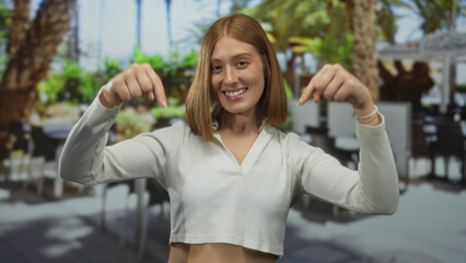 Young blonde woman points fingers downward at menu table on sunlit outdoor cafe terrace; invitation engagement focus attention.