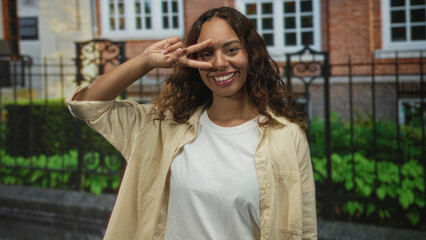 Woman smiling and holding a v sign with two fingers to her eye in front of building entrance outdoors, wearing white t shirt and beige overshirt; joy.