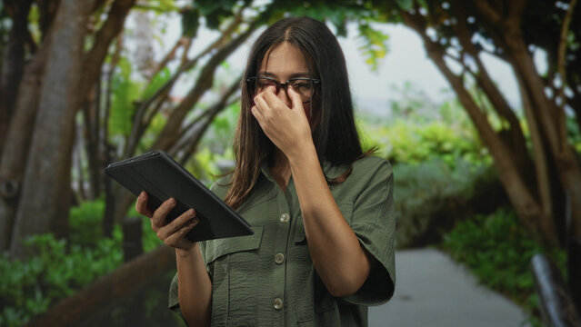 Woman pinching nose and holding tablet on forest path under trees, wearing glasses and green shirt; disgust.