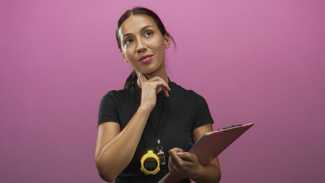 Young hispanic woman trainer holding clipboard and stopwatch with whistle necklace, touching chin in pink studio amid soft lighting; thoughtful planning.