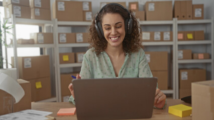 Woman wearing headphones smiling and gesturing with pen at laptop amid parcels on shelves in...