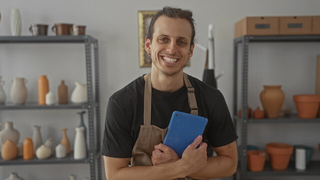 Man holding tablet to chest in pottery studio among shelves of handmade ceramics, vases and terracotta pots wearing an apron and smiling; craft pride.