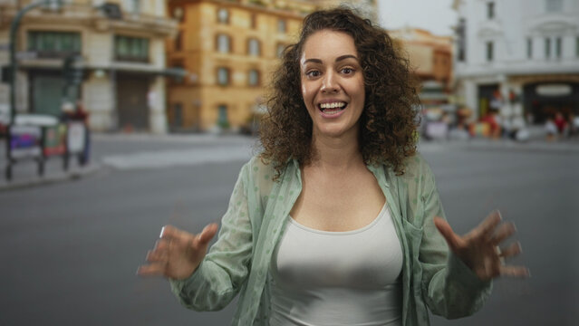 Young hispanic woman raises hands and smiles wide on a city street wearing white tank top and rings; joyful surprise. - Powered by Adobe