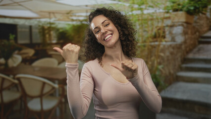 Woman smiling and showing thumbs up in a pink top on a restaurant terrace beside a stone building; casual joy.