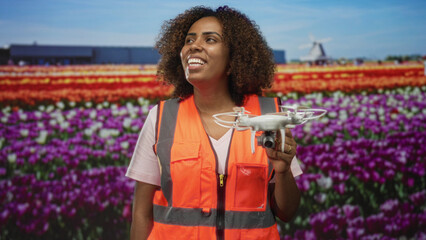 Young african american woman holds a quadcopter drone in hand beside rows of tulips in front of a building at a flower field; joy innovation.