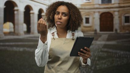 Woman baker wearing apron holds tablet and gestures with hand in old town building courtyard, eyes...