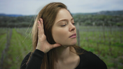 Obraz premium Woman cups ear with hand and closes eyes among green vineyard rows under cloudy sky in forest; curiosity.