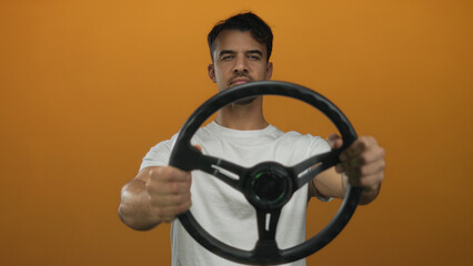 Young hispanic man holding a steering wheel looks worried against an isolated orange background, conveying a sense of stress or concentration.