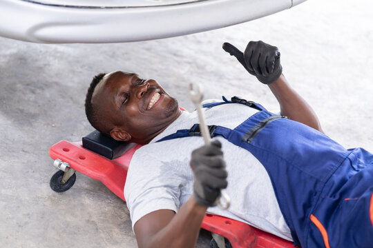 Happy African American Mechanic Working Under Car Giving Thumbs Up,Cheerful Automotive Technician Lying on Creeper Smiling and Holding Wrench.