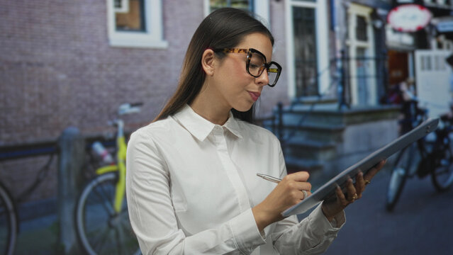 Woman writing on clipboard in street wearing white shirt and tortoiseshell glasses while holding pen; concentration planning.