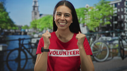 Woman smiling and clenching fists in red volunteer tshirt on a street beside an amsterdam canal; community service joy.