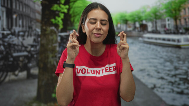 Young woman with fingers crossed on a street by a canal in amsterdam wearing a volunteer shirt; hope community support.