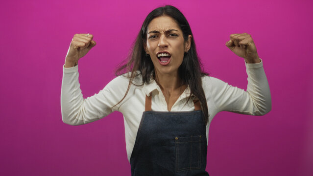 Young hispanic woman wearing denim apron flexes both fists showing biceps in pink studio; confidence empowerment strength.