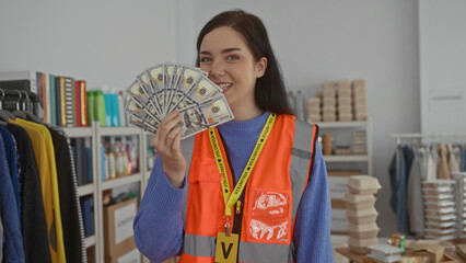 Woman wearing a volunteer vest indoors holds us dollars in a room filled with supplies suggesting charity efforts.