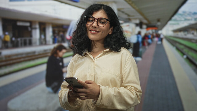 Woman holding smartphone, hands typing, standing on a train platform inside a station building with rails visible; calm focus.