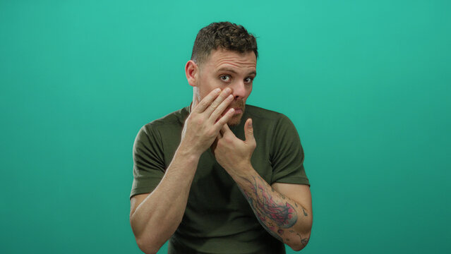 Young man with tattoos touching face against an isolated green background wearing casual clothing with concerned expression showcasing personal care and skincare awareness