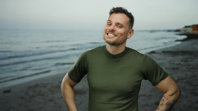 Young man with tattoos smiling confidently while standing on a serene beach during sunset, with ocean waves gently lapping in the background under a clear sky. - Powered by Adobe