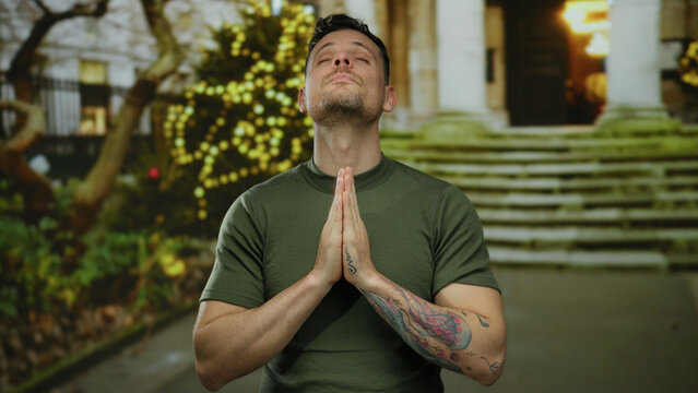 Young man in green shirt stands on urban street with hands in prayer gesture, lights glowing in background, serene expression, tattoo on arm, evening time, peaceful pose outdoors.