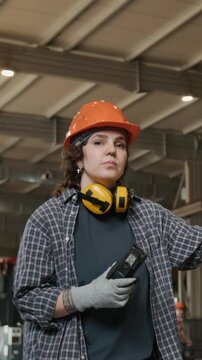 Vertical portrait shot of young forewoman in orange hard hat holding walkie talkie while directing heavy lifts in metalworking factory, slow motion