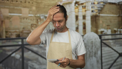 Man with blond hair and apron holds notebook and touches forehead in building among ancient ruins;...