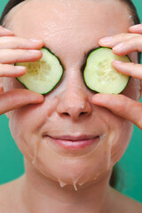 Portrait of a young woman with a fabric cosmetic mask on her face and a cucumber mask on her eyes on a green background. Self-care concept at home. Vertical photo