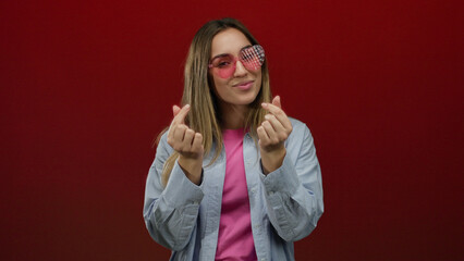 Woman making heart gesture wearing heart-shaped glasses with confident smile over red background isolated joyful female fashion pop art concept