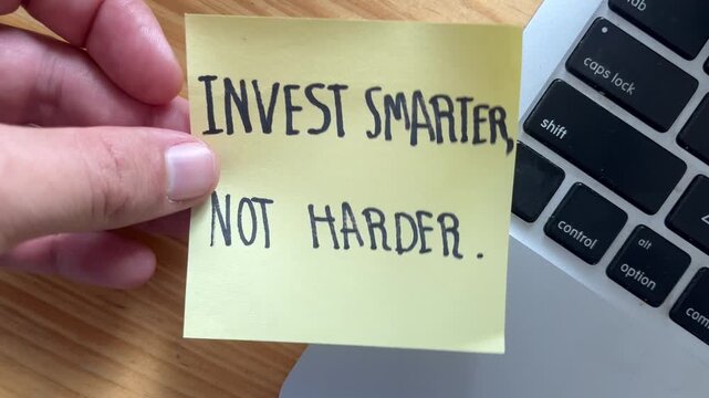 Hand Holding Yellow Sticky Note with ""Invest Smarter, Not Harder"" Message Near Laptop Keyboard