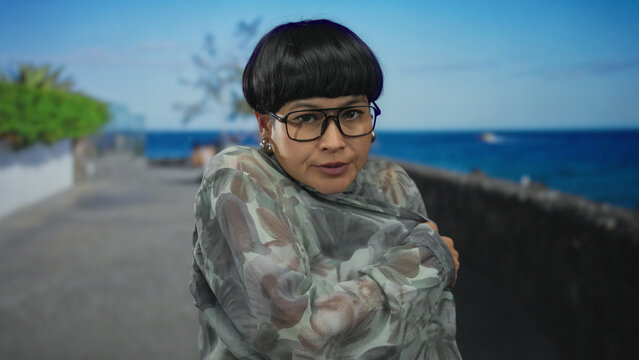 Woman shivering in cold on seaside promenade with ocean view wearing glasses and stylish outfit in an outdoor setting capturing winter chill and relentless wind.
