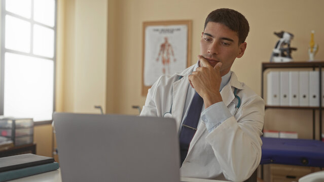 Man doctor with stethoscope holds chin on hand while reviewing medical data on laptop in hospital building; contemplation.