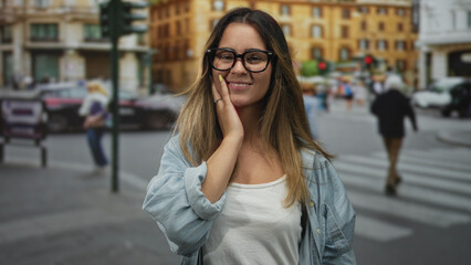 Fototapeta premium Young hispanic woman wearing round glasses and blue shirt smiles with hand on chest at busy city street crosswalk; gratitude serenity.