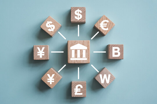 Wooden blocks displaying global currencies like dollar euro yen pound bitcoin and won connected to a central bank symbol representing international finance and exchange rates - Powered by Adobe