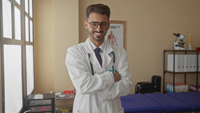 Man with stethoscope crossing arms in bright clinic wearing white coat and tie; professional confidence.