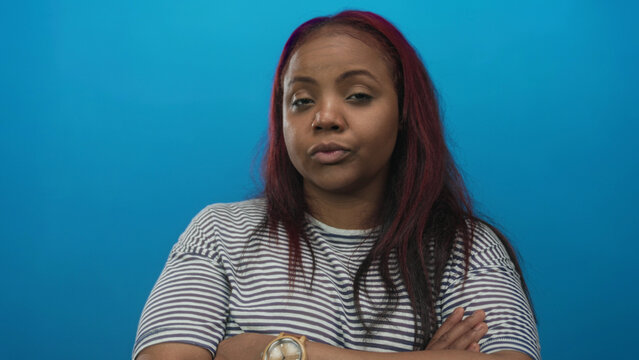 Black woman with arms crossed in striped shirt looks unimpressed with neutral gaze in blue studio setting; rejection.