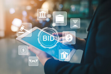 Person using a digital tablet displaying "BID" surrounded by auction and finance icons representing online bidding and financial transactions in a modern technologydriven environment