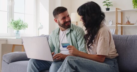 Couple with laptop and credit card making online shopping at home. Happy young people sitting on sofa together, smiling woman showing card while man planning money transaction, safe easy payment - Powered by Adobe