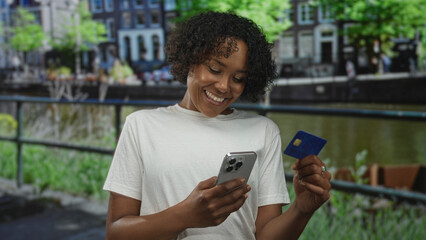 Woman holds smartphone and creditcard by a canal railing while smiling broadly on a city street; confidence.