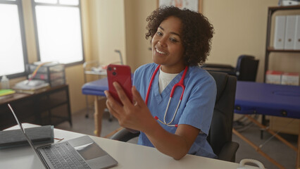 Woman doctor in blue scrubs holds red smartphone and touches chest while seated at desk with laptop in clinic; empathy trust.