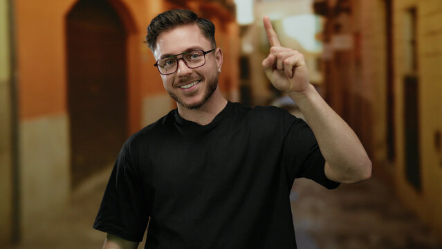 Young man with beard cheerfully pointing upward on a vibrant city street, exuding confidence and engaging charm in casual attire amid urban backdrop. - Powered by Adobe