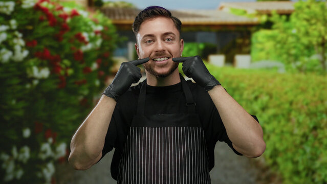 Young man with beard in apron and gloves smiling outdoors on a city street, posing with greenery background and vibrant floral display, conveying hospitality and friendliness.