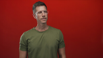 Young caucasian man in olive green t shirt with clear glasses looking sideways in studio with red...