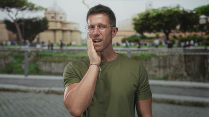 Man wearing glasses and green t shirt with hand on cheek in street by stone steps and trees;...