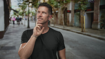 Fototapeta premium Man, young caucasian, on city street with hand on chin in thinking pose, wearing black tshirt and bracelet; contemplation.