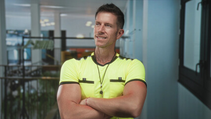 Referee man in neon yellow jersey with whistle on lanyard, smiling with arms crossed and bare forearms visible in hotel building; confidence.