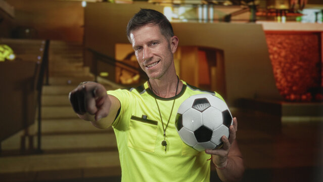 Young man wearing yellow shirt holding soccer ball and points finger toward lens in building lobby by staircase; confidence team encouragement.